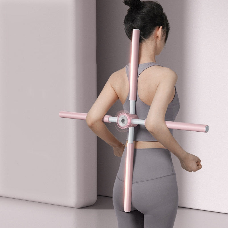 A woman in gray workout clothes uses a pink Yoga Stick with crossed bars on her back, standing in a minimalist room.